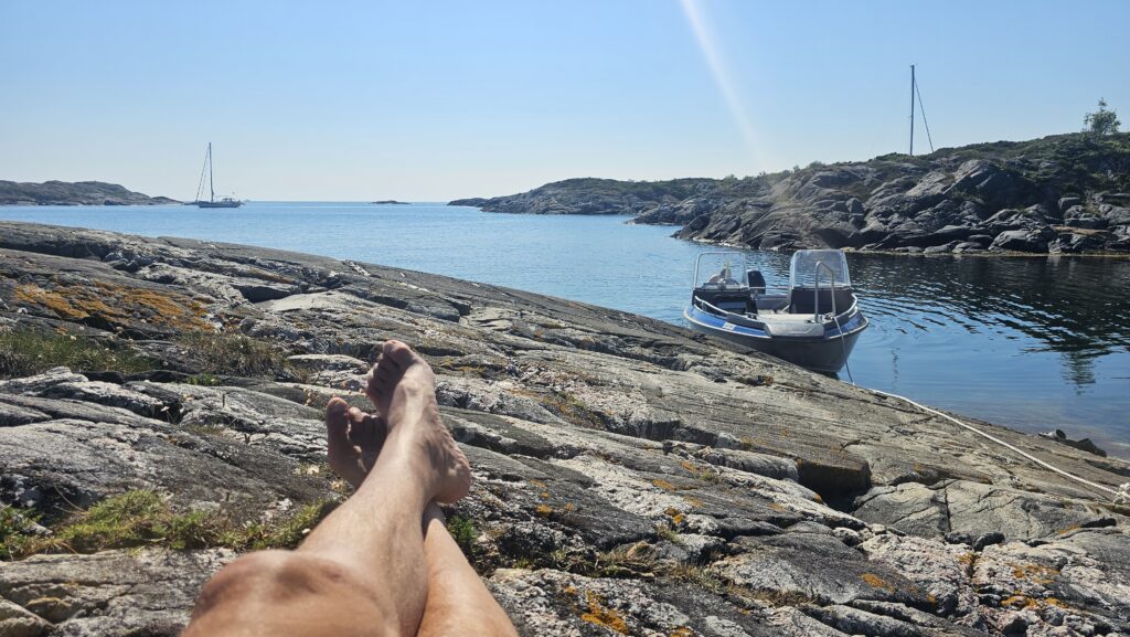 Avslapping i sola ved kysten i Sørlandet – beina i været, blikket mot havet, og båten klar for nye turer./ Speed Motorcenter / SpeedMC / Kyrre Hagen / MC-tur / MC-ferie / Motorsykkel / MC